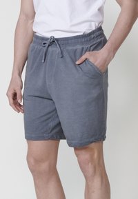 Uomo con pantaloni corti blu a trama waffle e una maglietta bianca, con la mano appoggiata nella tasca destra, in piedi contro uno sfondo chiaro.