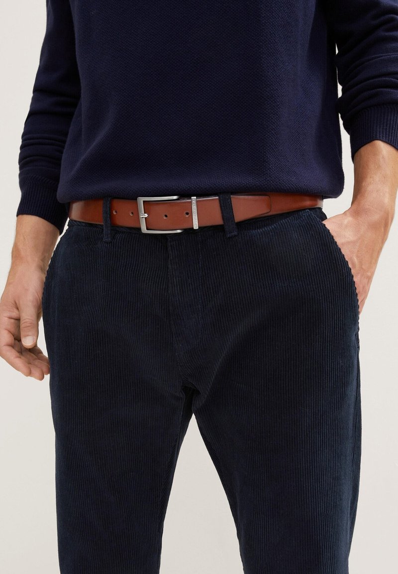 Pantalons en velours côtelé bleu marine avec des côtes verticales, associés à une ceinture en cuir marron dotée d'une boucle rectangulaire en argent et de détails de couture subtils.