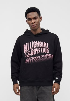 Jeune homme portant un sweat à capuche noir avec un grand texte rose "Billionaire Boys Club" et un graphisme de casque d'astronaute sur fond uni.