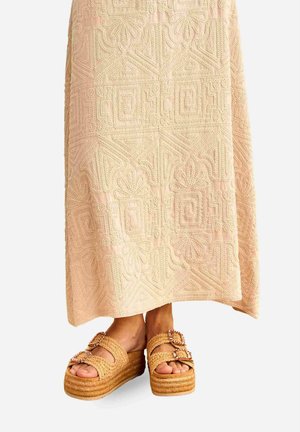 Pies que llevan sandalias de plataforma tejidas con correas de doble hebilla, parcialmente cubiertos por una falda larga beige con patrones geométricos texturizados.