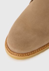 Botină din suede maro, cu vârf rotund, cusături decorative de-a lungul talpii și un bază din cauciuc de culoare deschisă.