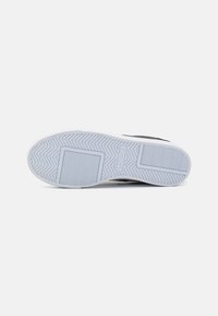 Suela de zapatilla negra con una parte inferior de goma blanca, presenta un patrón de espiga y detalles del logo. Textura suave con bordes redondeados.