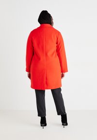 Rød ullkåpe med en strukturert krage og lange ermer, kombinert med mørke bukser og svarte hælboots. Enkel, ren linjer uten pynt.