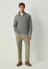 Pulover gri cu fermoar, cu mâneci lungi și textură cu grinzi, pantaloni bej și pantofi maro cu șireturi. Modelul stă cu mâinile în buzunare.
