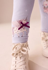 Levanduľové legíny s motívom jednorožca, ružovými akcentmi a fialovou mašľou. Kombinované s bielymi vysokými teniskami.