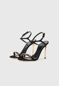 Black suede high-heeled sandals with adjustable ankle straps. Features a gold-tone chain accent and a stiletto heel design.