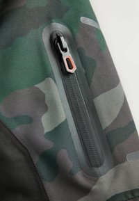 Green and black camouflage fabric featuring a horizontal pocket with a black zipper and an orange accent on the zipper pull. Texture visible.