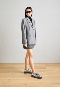 Grijs wollen blazer over een zwarte coltrui, gecombineerd met een gestreepte zwart-wit minirok en zilveren loafers met zwarte zolen. Zwarte zonnebril.