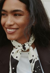 White and black polka dot scarf tied around the neck, featuring a smooth texture and a contrasting dark accent. Worn with a dark blazer.