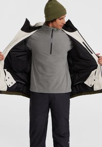 Manteau isolé pour homme avec un extérieur blanc, des accents noirs et un intérieur vert. Chemise en polaire grise à fermeture éclair en dessous. Pantalon noir.