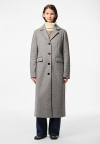 Mladá žena stojí, má na sobě dlouhý šedý kabát na knoflíky, krémový rolák, tmavě modré džíny a černé boty na bílém pozadí.