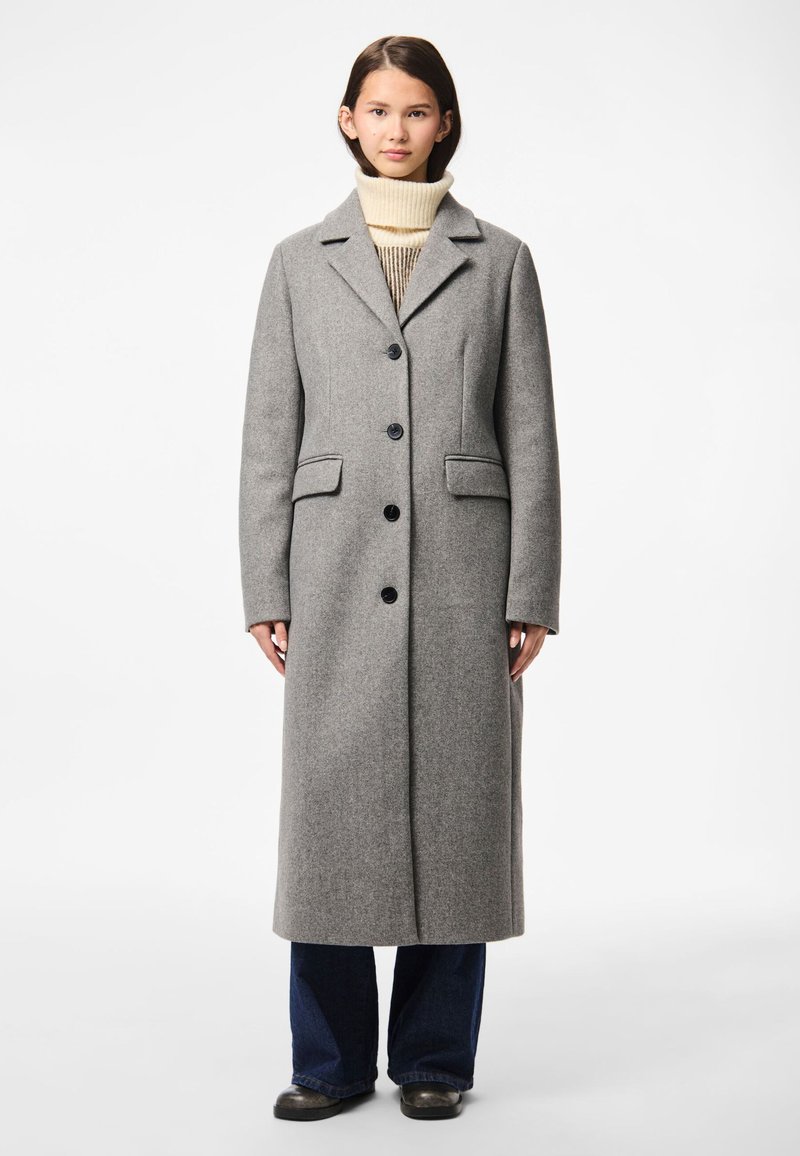 Mladá žena stojí, má na sobě dlouhý šedý kabát na knoflíky, krémový rolák, tmavě modré džíny a černé boty na bílém pozadí.