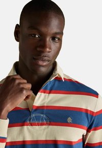 Nuori mies, joka pukeutunut kerma-, punainen- ja siniraidalliseen rugby-paitaan, pitää kaulusta oikealla kädellään ja katsoo mietteliäänä eteenpäin.