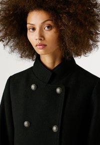 Μαύρο μάλλινο παλτό με ψηλό γιακά, σχέδιο διπλού κουμπώματος και μεταλλικά κουμπιά σε ασημί απόχρωση. Υφασμάτινη επιφάνεια με κομψή εφαρμογή.