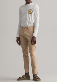 Chemise blanche à manches longues avec logo rond, pantalon beige et sandales vertes à enfiler. Accent sur des lignes épurées et une coupe décontractée. Sans motifs ni textures.