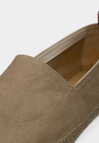 Μπεζ πλεκτό παπούτσι τύπου slip-on με ραμμένες λεπτομέρειες και πλεγμένη άκρη σόλας, παρουσιασμένο πάνω σε λευκό φόντο.