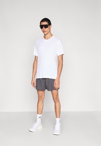 Valkoinen urheilupaita, jossa pieni sininen logo, yhdistetty harmaisiin shortseihin. Asusteina mustat aurinkolasit ja valkoiset lenkkarit.