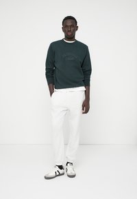 Tmavě zelená mikina se nápisem "ORIGINALS", spárovaná se světle šedými tepláky. Model má na sobě černobílé tenisky a stojí na bílém pozadí.