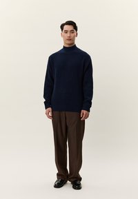 Mladi moški stoji, oblečen v temno moder teksturiran pulover, rjave hlače in črne polirane čevlje, na svetli enobarvni podlagi.