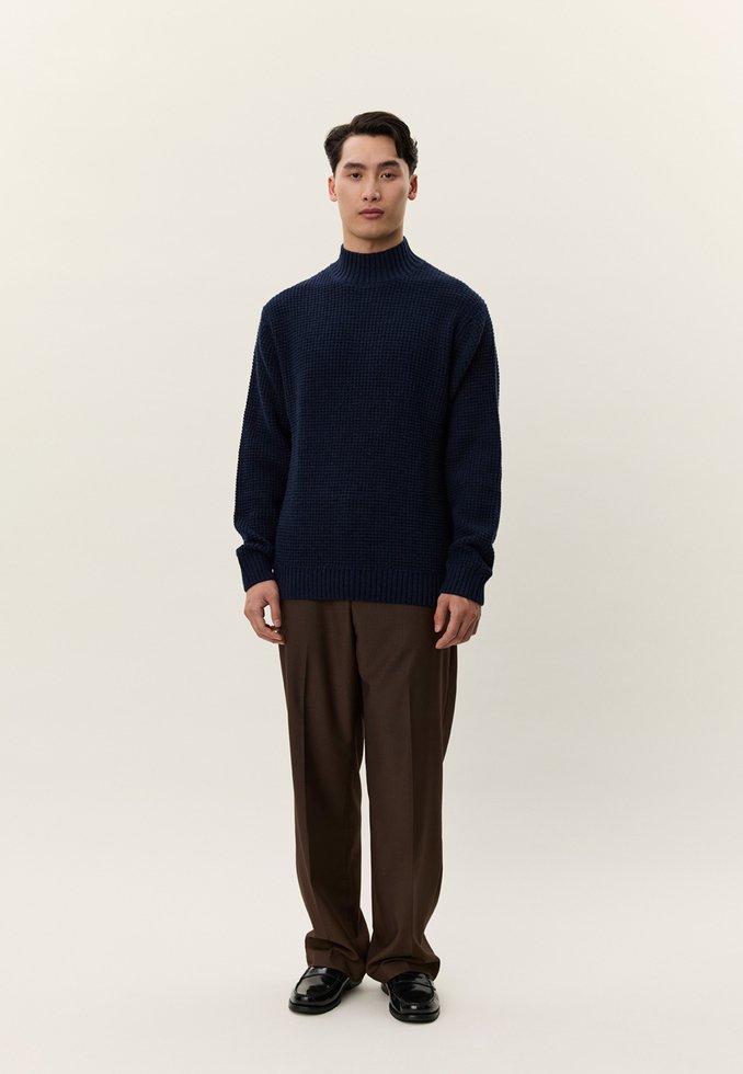 Mladi moški stoji, oblečen v temno moder teksturiran pulover, rjave hlače in črne polirane čevlje, na svetli enobarvni podlagi.