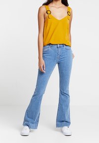 Donna che indossa un top senza maniche giallo senape con cinturini a fibbia, jeans svasati blu chiaro e sneakers bianche, in piedi su uno sfondo bianco.