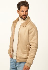 Beige Bomberjacke mit strukturiertem, künstlichem Lammfellkragen, Reißverschluss und seitlichen Taschen. Getragen über einem hellen Rollkragensweater.