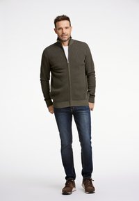 Olivgrön zip-up tröja med en texturerad mönster, bärs över en vit skjorta, ihop med blå jeans och bruna sneakers.
