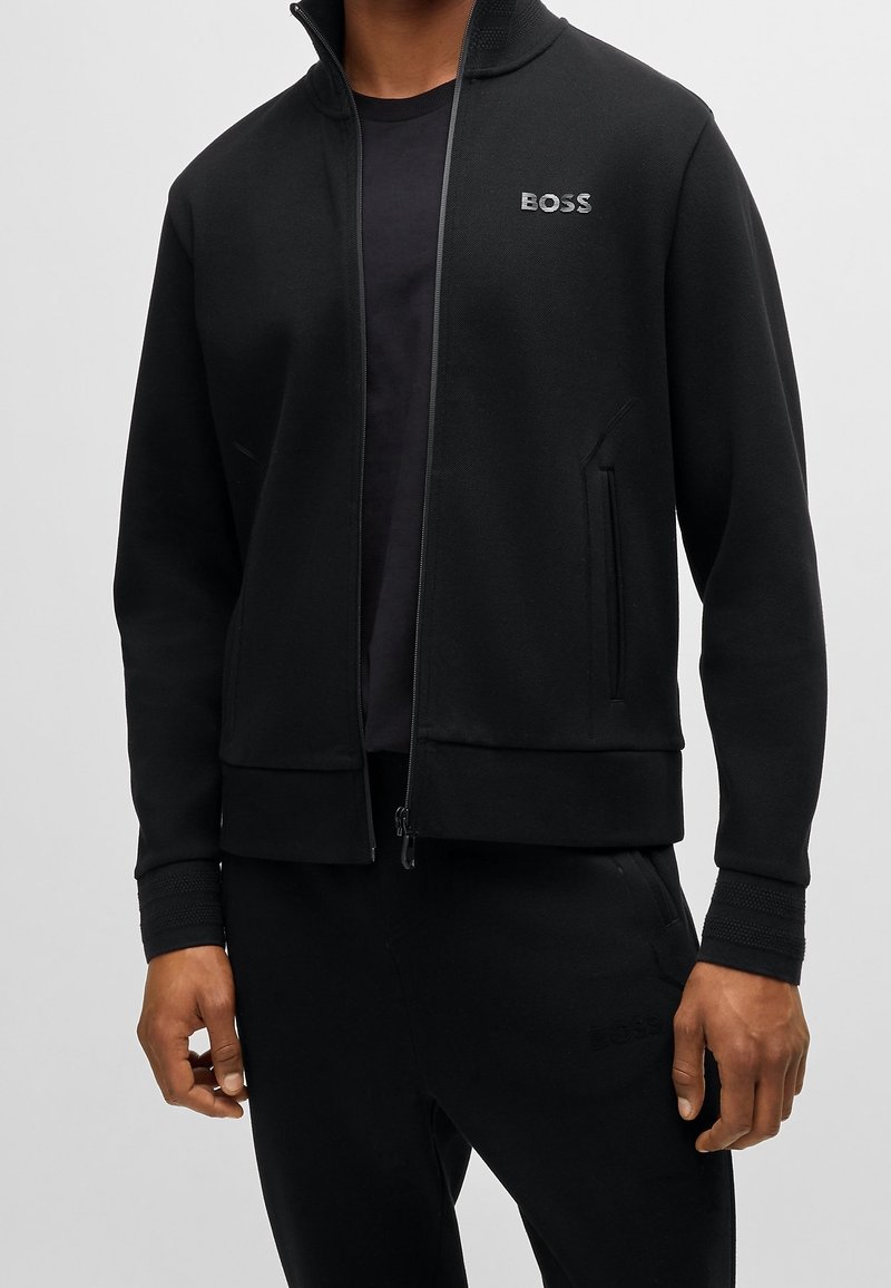 Homme portant une veste zippée noire avec le logo BOSS sur la poitrine, associée à un pantalon noir assorti sur un fond clair uni.