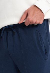 Marinblå joggers i ett mjukt tyg med elastisk midja, dragsko och sidofickor. Sömnadsdetaljer längs sömmarna.