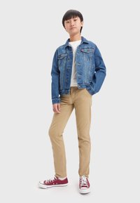 Giacca di jeans blu con due tasche sul petto, t-shirt bianca, pantaloni khaki e sneakers rosse con suola e lacci bianchi.