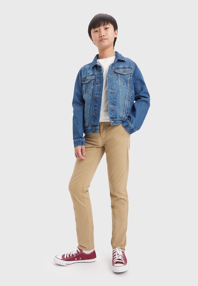Giacca di jeans blu con due tasche sul petto, t-shirt bianca, pantaloni khaki e sneakers rosse con suola e lacci bianchi.