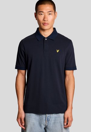 Jonge man die een marineblauw poloshirt draagt met een geel adelaarlogo op de borst en lichtblauwe spijkerbroek, staand tegen een effen grijze achtergrond.