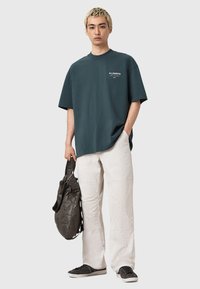 Jeune homme aux cheveux blonds portant un t-shirt noir surdimensionné, un pantalon clair, des baskets noires, tenant un sac en cuir matelassé noir contre un fond neutre.