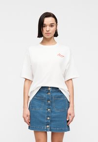 Γυναίκα με κοντά σκούρα μαλλιά που φοράει ένα λευκό φαρδύ t-shirt με κόκκινο κείμενο και μια μπλε τζιν φούστα με κουμπιά και μπροστινές τσέπες.