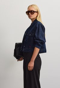Donna con i capelli biondi lunghi indossa occhiali da sole scuri, una giacca di jeans blu scuro, pantaloni neri e tiene una borsa nera in mano.