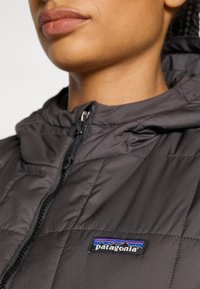 Juodas izoliuotas švarkas su pūkuotu dizainu, su užtrauktuku ir gobtuvu. Ant krūtinės yra ryškus Patagonia logotipas.