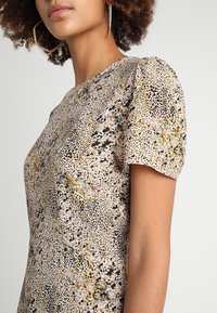 Robe à manches courtes en tissu imprimé avec des points et des éclaboussures dorés, verts et noirs sur un fond rose clair. Texture lisse.