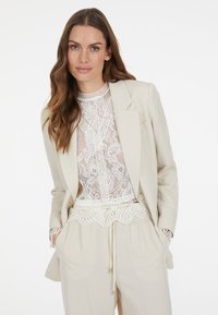 Leinenblazer in hellem Beige, kombiniert mit einem weißen Spitzentop mit aufwändigen Blumenmustern und einem Kordelzugbund für eine legere Passform.