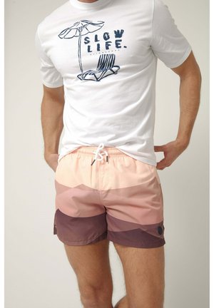 Hombre con camiseta blanca con el lema "Slow Life" y un diseño de silla de playa, combinado con pantalones cortos bicolor rosa y marrón, manos en los bolsillos.