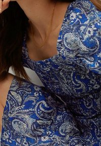 Frau in einem blauen gemusterten Kleid mit weißen Blumen- und Paisleymustern, accessoriert mit einer dünnen goldenen Kettenhalskette.
