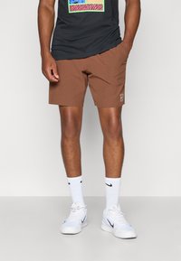 Brune shorts med et enkelt design, sidelommer og en liten hvit logo. Kombinert med en svart t-skjorte og hvite sneakers med svarte detaljer.