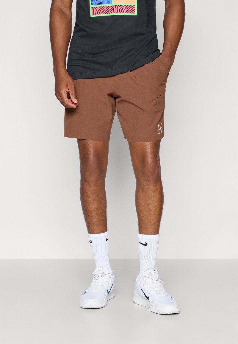 Brune shorts med et enkelt design, sidelommer og en liten hvit logo. Kombinert med en svart t-skjorte og hvite sneakers med svarte detaljer.