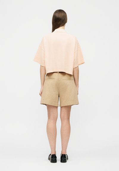 Γυναίκα που φοράει ένα κοντό ροζ ανοιχτόχρωμο μπλουζάκι με κοντά μανίκια, μπεζ σορτς με ψηλή μέση και μαύρα μοκασίνια, εμφανιζόμενη από πίσω με φόντο λευκό.