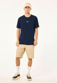 Navy short-sleeve T-shirt with a logo, paired with beige shorts. The model wears beige sneakers and white socks with a logo. Casual outfit design.
