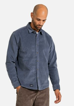 Mann mit rasiertem Kopf trägt ein dunkelblaues, langärmeliges Hemd mit Knöpfen und braune Hose, schaut nach unten mit einer Hand in der Tasche.