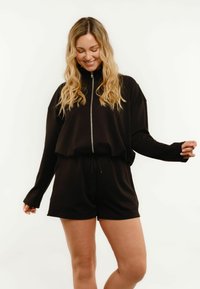 Femme aux longs cheveux blonds, vêtue d'un haut noir à manches longues zippé et d'un short noir, souriante et regardant vers le bas avec une posture détendue.