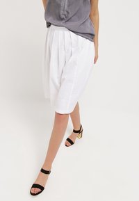 Kvinna klädd i vita veckade knälånga shorts, grå ärmlös topp och svarta klacksandaler framför en plain vit bakgrund.