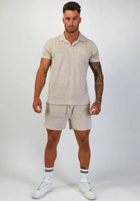 Beige bomullspolo med korta ärmar och bröstficka, kombinerad med matchande shorts. Modellen bär vita sneakers med svarta ränder på strumporna.