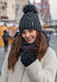 Bonnet en tricot gris avec un pompon, écharpe épaisse assortie et gants noirs. Tissu texturé, design côtelé avec une étiquette de logo visible.