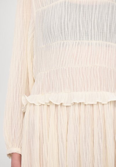 Lightweight, cream-colored dress with textured, crinkled fabric. Features ruffled hem and long sleeves. Vertical pleats create movement.